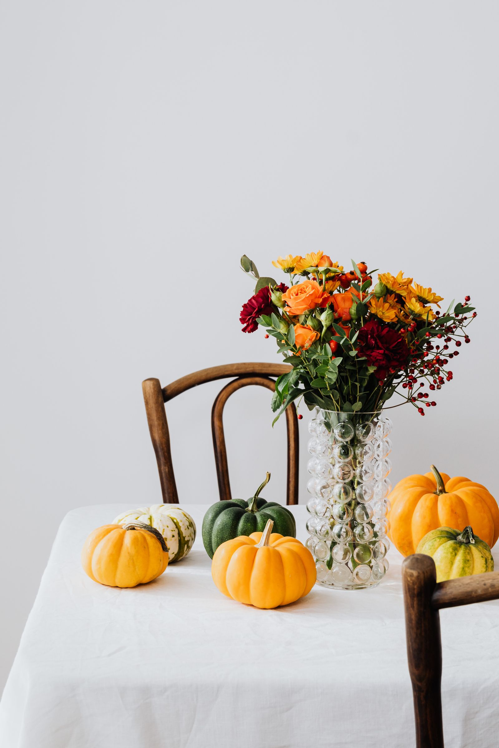 An autumn bouquet in orange and red in a glass vase beside small pumpkins