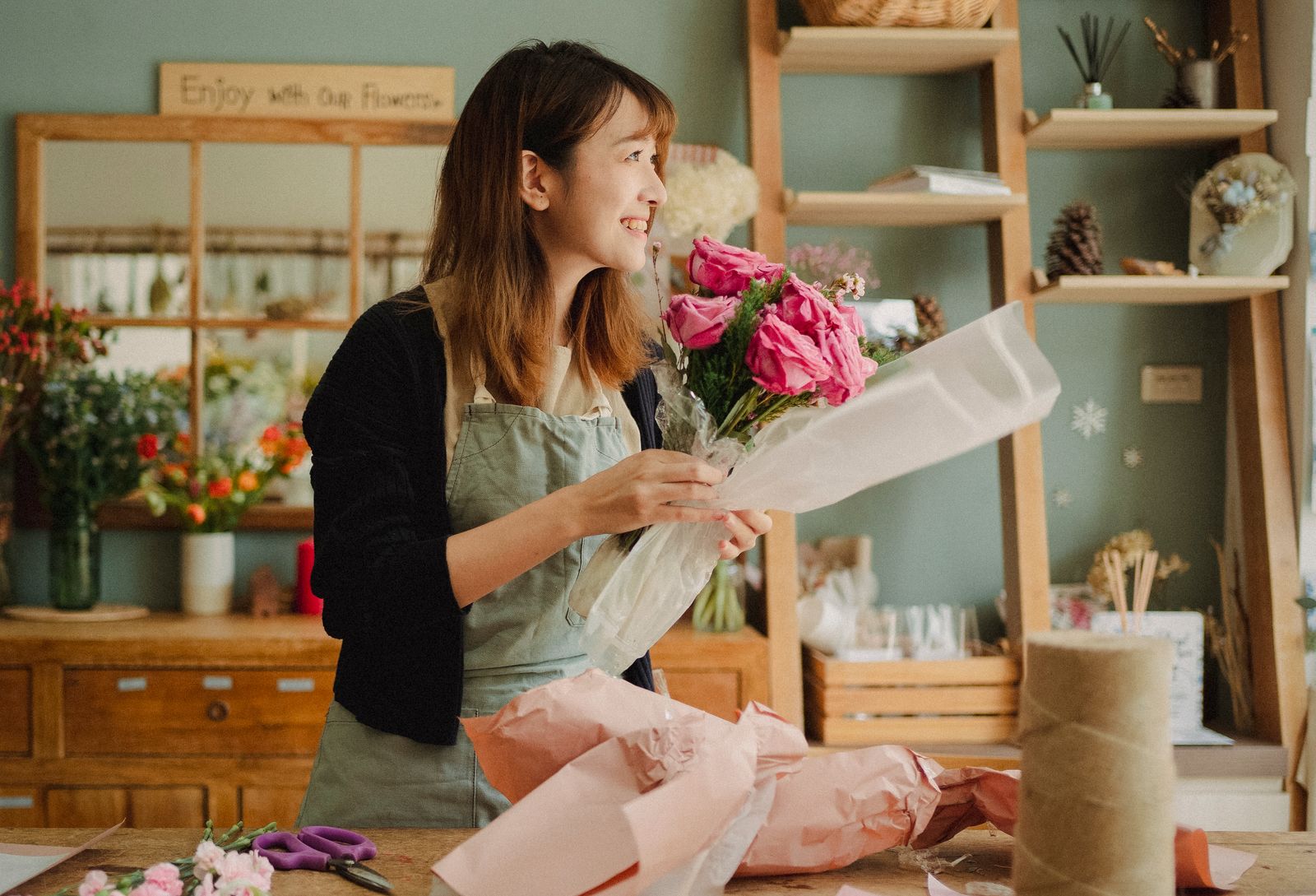 A florist in an apron wrapping a bouquet in pink paper at a workbench in a flower shop